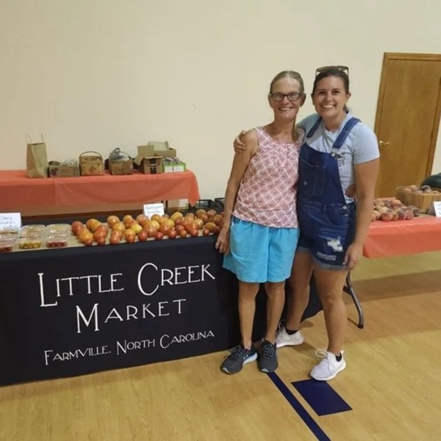 Little+Creek+Market+-+eatwithintention Two women pose together at the Little Creek Market booth in Farmville, North Carolina, showcasing fresh produce, including a variety of apples. The market table features a black tablecloth with the market's name and a selection of local goods in the background.